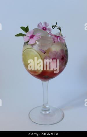 Eleganter Cocktail garniert mit rosa Blumen, einer Limettenscheibe und Minzblättern, serviert in einem Glas mit Eiswürfeln Stockfoto