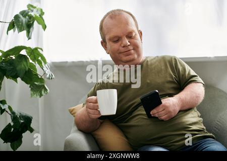 Ein Mann mit Inklusivität sitzt in einem bequemen Stuhl zu Hause und genießt eine Tasse Kaffee, während er auf sein Telefon schaut. Stockfoto