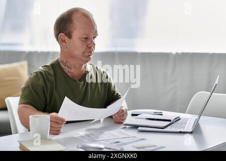 Ein Mann mit Inklusivität sitzt an seinem Schreibtisch zu Hause und liest sich Papiere, während ein Laptop in der Nähe sitzt. Stockfoto