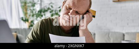 Ein Mann mit Inklusivität sitzt zu Hause, anscheinend von finanziellen Bedenken belastet, während er ein Dokument prüft und eine Kreditkarte besitzt. Stockfoto
