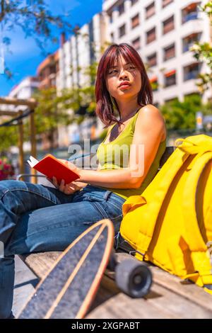 Vertikales Foto einer chinesischen jungen Frau, die Notizen auf einem Tagebuch schreibt und auf einer Parkbank neben dem Skateboard sitzt Stockfoto