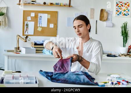 Ein junger Mann in einem weißen Hemd inspiziert ein lebendiges Denim-Kleidungsstück in seiner Werkstatt zur Bekleidungsrestaurierung. Stockfoto