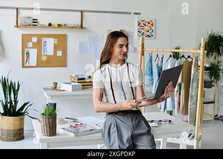 Ein junger Mann arbeitet an seinem Laptop in seinem Bekleidungsatelier, umgeben von lebendigen Stoffen und Pflanzen. Stockfoto