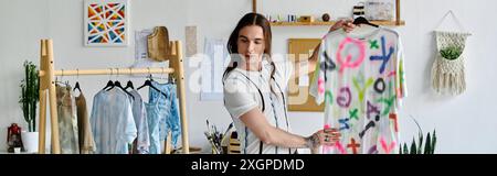 Ein junger Mann steht in seinem Atelier und zeigt stolz ein lebendiges, handgemaltes T-Shirt. Er setzt sich für nachhaltige Praktiken ein, indem er alten Menschen neues Leben einhaucht Stockfoto