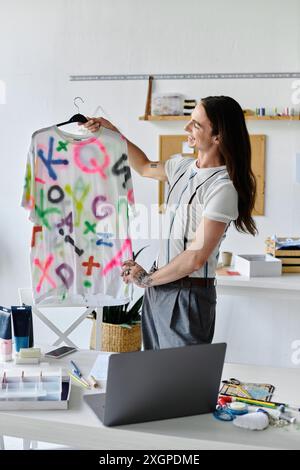 Ein junger Mann in einem Heimwerkeratelier zeigt stolz ein lebhaftes, aufgeputztes T-Shirt. Stockfoto