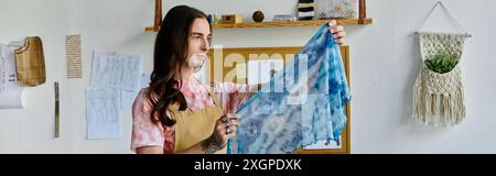 Ein junger Mann in einem hellrosafarbenen T-Shirt und einer Schürze hält einen leuchtend blau-weißen Stoff in seiner Bekleidungswerkstatt. Stockfoto