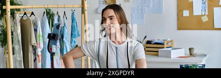 Ein junger Mann lächelt, während er in seinem Bekleidungsatelier arbeitet, umgeben von Regalen aus lebendigen Stoffen und einer Telefonkamera, um seine Kreationen festzuhalten Stockfoto