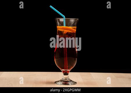 Ein Glas Eistee mit Zitronenscheiben und blauem Strohhalm auf einem Holztisch vor schwarzem Hintergrund Stockfoto