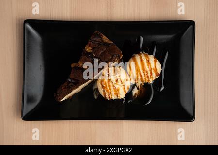 Ein Blick von oben auf ein köstliches Dessert mit einem Stück Schokoladenkuchen und zwei Kugeln Vanilleeis auf schwarzem Teller über Holztisch Stockfoto