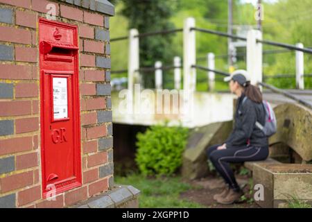 Rotes, Royal Mail Postfach/Briefkasten mit königlichem Chiffer GR in einer Backsteinmauer in einem traditionellen englischen Dorf auf dem Land, mit weiblichen Sitzen im Hintergrund. Stockfoto