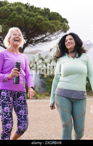 Zwei glückliche ältere Freundinnen laufen zum Sport auf der Landstraße, lachen, Kopierraum. Freundschaft, Fitness, Bewegung, Aktivität und Gesundheit Stockfoto