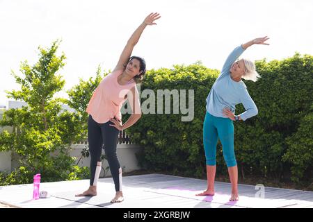 Zwei glückliche ältere Freundinnen üben Yoga im Stehen und Arm Strecken im sonnigen Garten. Yoga, Wohlbefinden, Bewegung, Freundschaft und Gesundheit Stockfoto