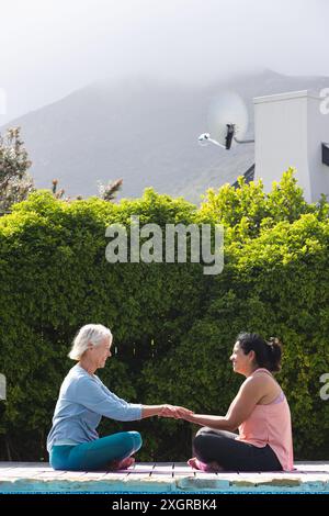 Zwei glückliche, verschiedene ältere Freundinnen üben Yoga, sitzen im sonnigen Garten, Kopierraum. Yoga, Wohlbefinden, Freundschaft und gesunder Seniorenstil, Stockfoto