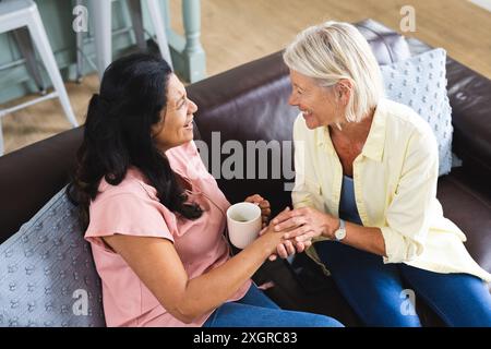 Zwei glückliche ältere Freundinnen, die Kaffee trinken, reden und Hände im Wohnzimmer halten. Freundschaft, Entspannung, Ruhestand, häusliches Leben und Stockfoto