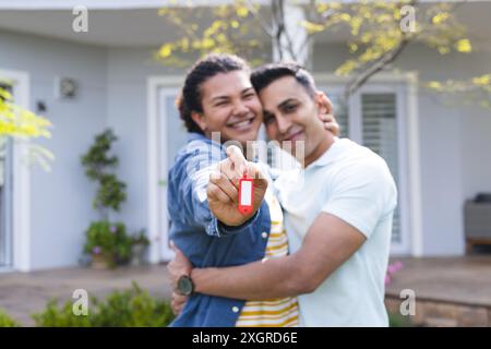 Diverse schwule Paare zeigen einen Schlüssel vor einem Haus. Sie zelebrieren Hausbesitz mit fröhlichen Ausdrücken in einer Wohngegend. Stockfoto