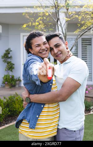 Diverse schwule Paare zeigen stolz Schlüssel zu einem neuen Zuhause, mit Kopierraum. Sie feiern gemeinsam Hausbesitz in einer sonnigen Umgebung im Freien. Stockfoto