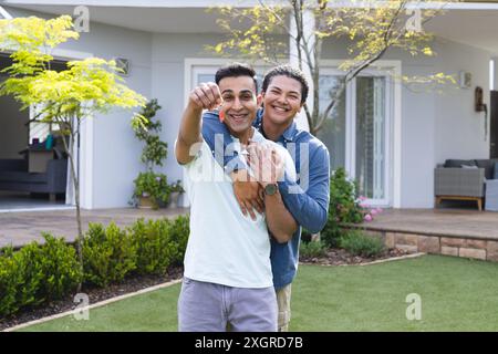 Diverse Paare zeigen Schlüssel vor einem neuen Zuhause. Sie zelebrieren Hausbesitz mit fröhlichen Ausdrücken in einer vorstädtischen Umgebung. Stockfoto