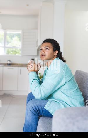 Ein junges birassisches schwules Paar sitzt nachdenklich auf einer Couch zu Hause. Sie wirken besinnlich, entspannend in einem hellen, luftigen Wohnbereich. Stockfoto
