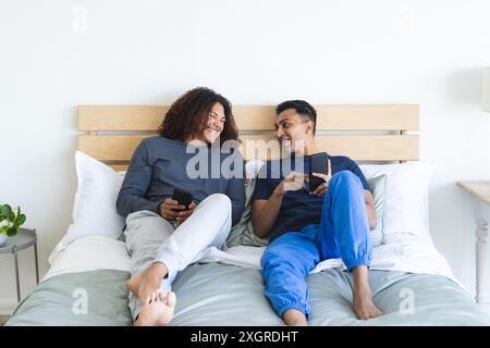 Schwules birassisches Paar entspannt sich in einem gemütlichen Schlafzimmer. Sie teilen einen Moment der Verbindung, während sie mit ihren Smartphones interagieren. Stockfoto