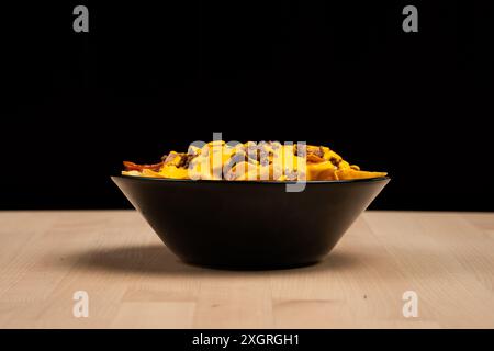 Eine Schüssel mit gefüllten Pommes frites, garniert mit cremiger Sauce und gegrilltem Huhn auf einem Holztisch vor schwarzem Hintergrund Stockfoto
