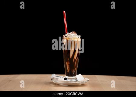 Ein köstlicher Schokoladen-Milchshake mit Schlagsahne und einem rot gestreiften Strohhalm auf Holztisch vor schwarzem Hintergrund Stockfoto
