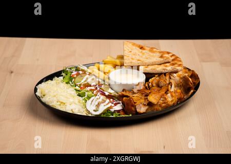 Ein köstlicher Teller gemischter Döner-Döner mit Salat, Pommes, Pita-Brot und Sauce auf einem Holztisch vor schwarzem Hintergrund Stockfoto