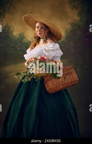 Porträt einer Frau in historischer Kleidung, in großem Strohhut, weißer Bluse und grünem Samtrock, hält Korbkorb mit viel Grün und Stockfoto