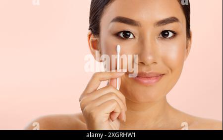 Junge Frau, die Wattestäbchen in der Nähe des Auges auf rosa Hintergrund benutzt Stockfoto