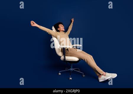 Foto in voller Länge des schönen jungen Mannes Sessel Sleepy Stretching gekleidetes stilvolles beiges Kleid isoliert auf dunkelblauem Hintergrund Stockfoto