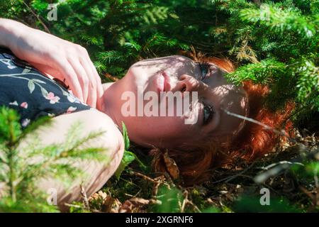 Frau mit rotem Haar liegt zwischen grünen Kiefernzweigen, Sonnenlicht wirft zarte Schatten auf ihr Gesicht. Sie blickt nach oben und strahlt eine ruhige und besinnliche Ausstrahlung aus Stockfoto