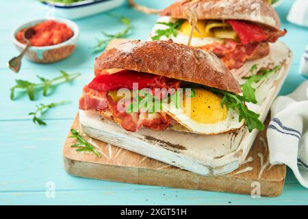 Sandwich-Lagen. Englisches oder amerikanisches frisch zubereitetes Picknick-Sandwich mit Ei, Rucola, Speck, Gurken und Tomaten auf einem türkisfarbenen Tisch Stockfoto