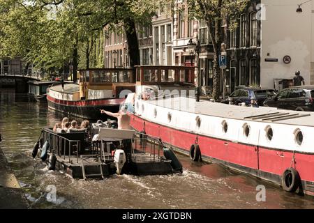 An einem sonnigen Tag in Amsterdam, den Niederlanden, fährt ein Boot voller Nachtschwärmer an Hausbooten vorbei, die an einem schmalen Kanal vor Anker liegen. Stockfoto