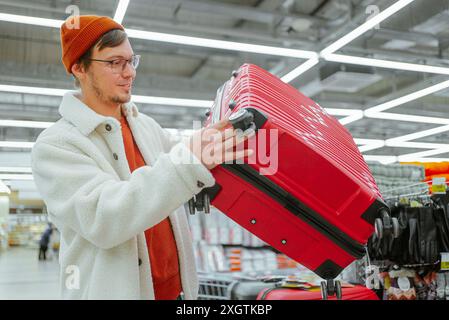Mann mit Mütze und Mantel untersucht den roten Koffer im Gang des Ladens und bereitet sich auf die Reise vor. Mann wählt Koffer im Supermarkt für Flugreisen. Stockfoto