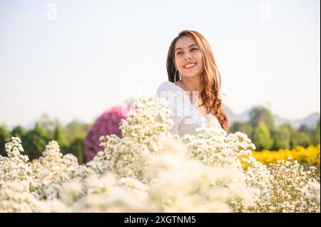 Glückliche schöne junge asiatische Frau, die auf weißen Schneideblumen steht und lächelt, die am sonnigen Tag im Garten blüht Stockfoto