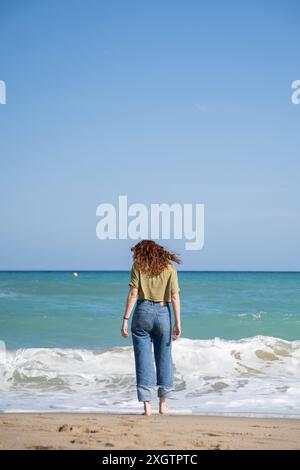 Rückansicht einer unerkennbaren Frau, die allein am Strand steht und zum Meer blickt, von hinten in einer ruhigen Szene festgehalten. Stockfoto