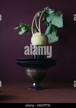 Ein elegant präsentierter Kohlrabi mit frischen Blättern, der auf einem kunstvollen Vintage-Metallkelch vor einem kräftigen kastanienbraunen Hintergrund thront und eine Mischung aus einem Mix aus Stockfoto