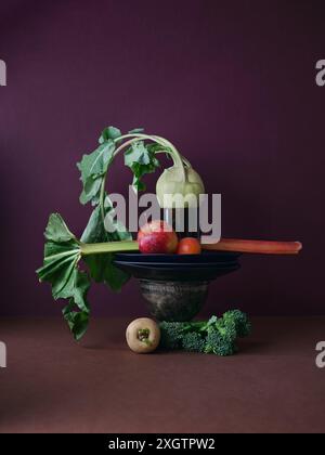 Eine kreative Präsentation von gesundem, frischem Gemüse, einschließlich Brokkoli, Kohlrabi, Äpfeln und einem belaubten Stiel, der auf einer Metallschale gegen ein tiefes braunes Maroo balanciert Stockfoto