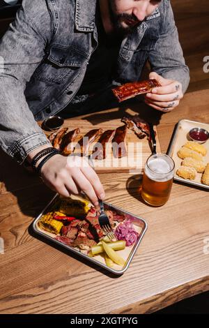 Ein tätowierter Mann sitzt an einem Holztisch und genießt verschiedene Grillgerichte wie Rippchen, Brustkorb, Mais und eingelegtes Gemüse, begleitet von einem Glas Stockfoto