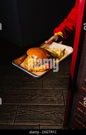 Eine Person in einer auffälligen roten Jacke serviert einen frisch zubereiteten Cheeseburger mit goldenen Pommes Frites auf einem Metalltablett. Das Bild fängt das warme Ambiente ein Stockfoto