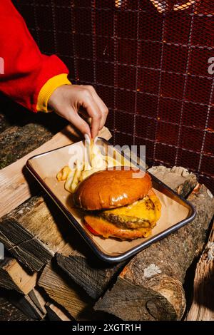 Eine Person schnappt sich einen Braten neben einem saftigen Burger mit geschmolzenem Käse und frischen Belägen, serviert auf einer rustikalen Holzplatte. Diese herzhafte Mahlzeit verkörpert ein gemütliches B Stockfoto