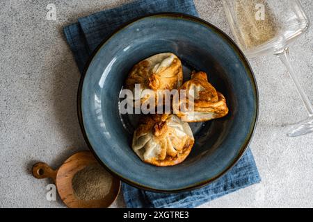 Eine Nahaufnahme traditioneller georgischer Khinkali-Knödel auf einer strukturierten blauen Keramikplatte, begleitet von einer hölzernen Schaufel mit Gewürzen und einem klaren Wein Stockfoto