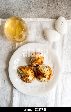 Eine rustikale Präsentation georgischer Khinkali, traditionelle Knödel, serviert auf einem weißen Teller neben einem Glas Weißwein und Salzstreuer, Symbol Stockfoto