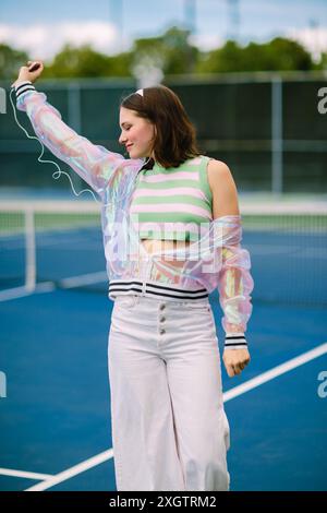 Eine junge Frau hört fröhlich Musik mit Kopfhörern und tanzt auf einem Tennisplatz. Sie trägt ein farbenfrohes, gestreiftes Oberteil und eine transparente Jacke Stockfoto