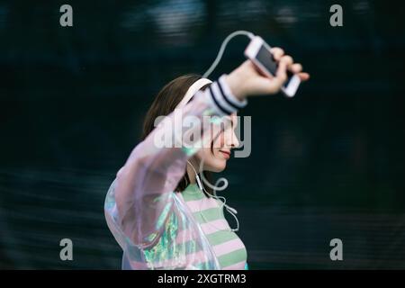 Eine junge Frau wird gefangen genommen, während sie Musik hört, indem sie weiße Kopfhörer verwendet, die mit einem Smartphone verbunden sind Stockfoto