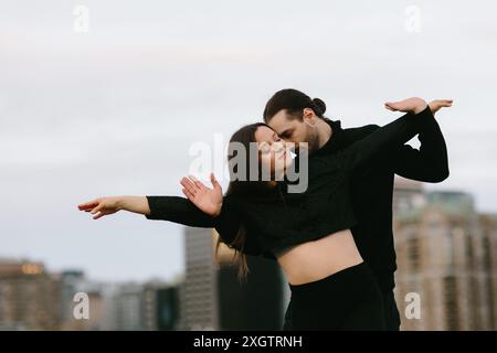 Ein junger Mann und eine junge Frau teilen einen zärtlichen Moment, während sie auf einem Dach der Stadt tanzen. Beide sind schwarz gekleidet und eng umarmen, mit dem Mann, der aussieht Stockfoto