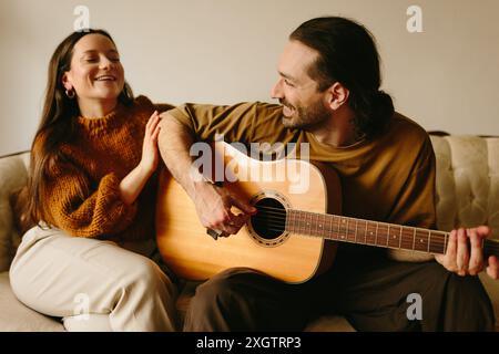 In einer fröhlichen Szene spielt ein Mann eine akustische Gitarre für eine lächelnde Frau, die auf einer Couch sitzt und einen Moment musikalischer Verbindung und Glück teilt Stockfoto