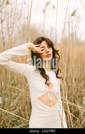 Eine junge Frau in einem stilvollen weißen Kleid posiert zwischen hohem, trockenem Gras, ihre Hand bedeckt anmutig einen Teil ihres Gesichts und schafft eine künstlerische Stimmung in einer Natur Stockfoto