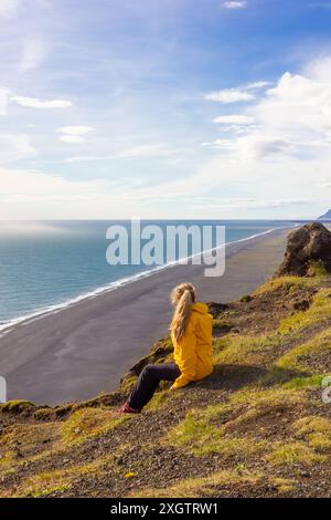 Eine besinnliche Frau sitzt in einer hellgelben Jacke auf einer Klippe und beobachtet den weiten, ruhigen schwarzen Sandstrand und das glitzernde Meer unter einem breiten Blau Stockfoto