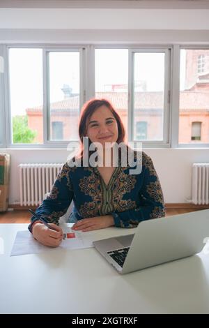 Eine fröhliche junge Geschäftsfrau sitzt an einem weißen Schreibtisch in einem hellen Büro und arbeitet an einem Laptop, während sie Stift und Papiere hält Stockfoto