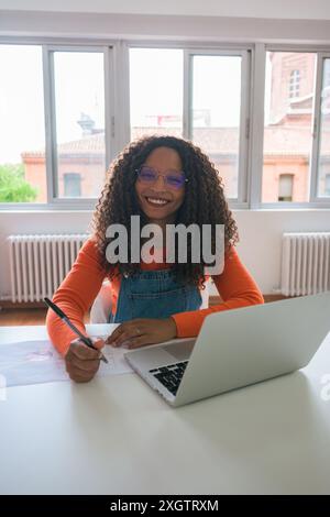 Eine schwarze Frau mit lockigen Haaren, die eine Brille und ein leuchtend orangefarbenes Hemd trägt, arbeitet fröhlich an ihrem Laptop an einem modernen weißen Schreibtisch in einem gut beleuchteten Licht Stockfoto